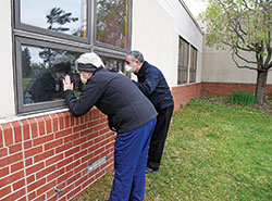 Therese Brandon, left, and Father Joseph Moriarty visit their father, Patrick Moriarty, on April 30 through the window of their father’s room at the St. Paul Hermitage retirement community in Beech Grove. For more than two months, visitors have not been allowed except for end-of-life situations to help protect the residents and staff members from the coronavirus. (Photo by Sean Gallagher)